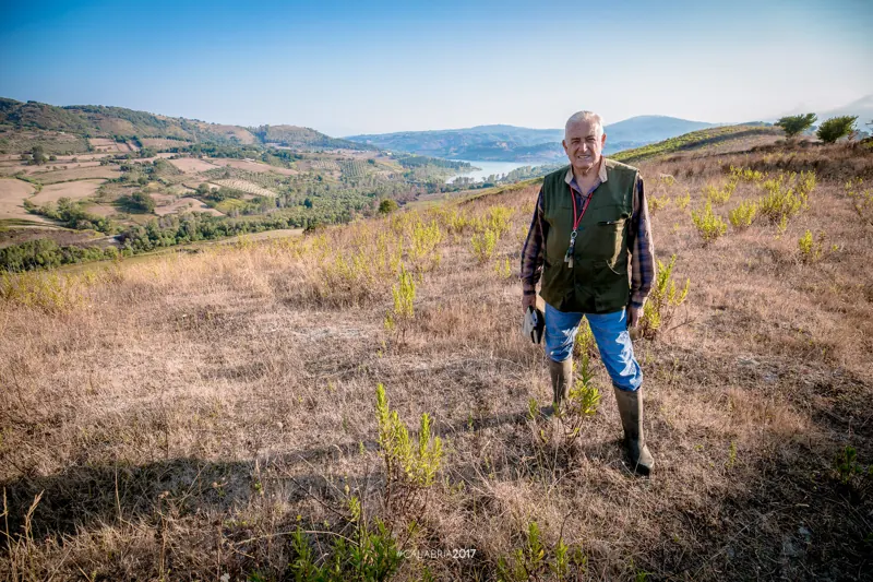 Aldo Geremicca in piedi su una collina d’erba secca, con una valle e un lago alle spalle